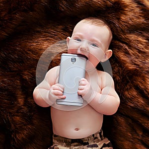 Funny happy baby boy gnaws a phone in his hands on a fur carpet. Smiling child holds a smartphone, top view