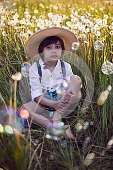 funny happy a beautiful boy child sit in hat on a field with white dandelions at sunset in summer. soap bubbles are