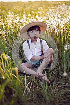 funny happy a beautiful boy child sit in hat on a field with white dandelions at sunset in summer. soap bubbles are