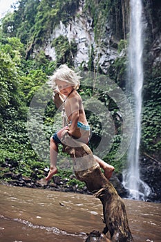 Funny child sit on snag under waterfall in tropical jungle