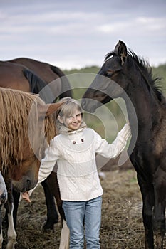 Funny happy child in a white sweater and jeans standing among horses foals on the farm smiling. Lifestyle portrait