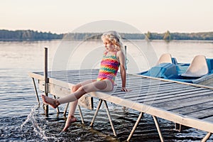 Funny happy cute Caucasian blonde girl child sitting on wooden deck pier by lake. Smiling laughing kid in swimsuit splashing with