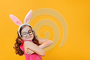 Funny happy cute little girl wearing Easter bunny ears and glasses on a yellow background. The child hugs himself with his arms