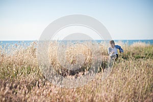 Funny happy healthy small caucasian kid running away among dry grass with blue sky and sea horizon on background during outdoor
