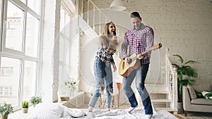 Funny happy and loving couple dance on bed singing with tv controller and playing guitar. Man and woman have fun during