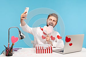 Funny happy man with beard in white shirt with red sticky hearts making selfie with pink heart and striped giftbox sitting at