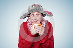 Funny happy man in a red sweater, scarf and winter hat holds a mug of hot drink in his hands