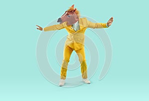 Funny happy man in yellow suit and horse mask dancing on turquoise studio background