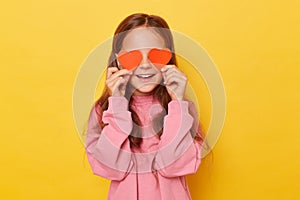 Funny happy smiling little girl wearing pink sweatshirt covering her eyes with red little hearts isolated over yellow background