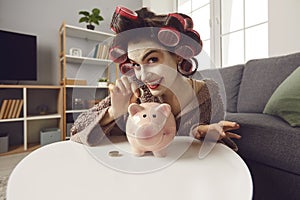 Smiling young woman with face mask and hair curlers putting coins in her piggy bank