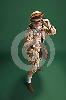 Funny image of young man in image of detective looking in magnifying glass against green studio background. Big teeth