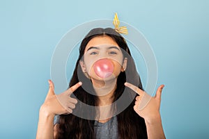 Funny Indian teen girl blowing bubble from sugar free chewing gum, pointing at it over blue studio background