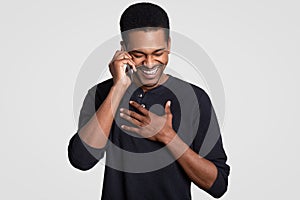 Funny joyful dark skinned masculine laughs happily, hears comic story from friend on cell phone, keeps hands on chest, giggles