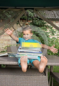 Funny joyful inquisitive preschooler sits on table, holding a large pile of books