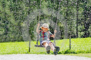 Funny kid boy having fun with chain swing on outdoor playground while being wet splashed with water. child swinging on