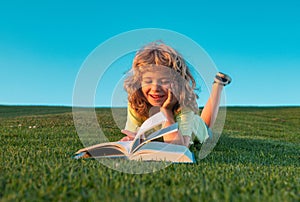 Funny kid reading the book in spring park. Child boy reading book, laying on grass in field on sky background. Portrait