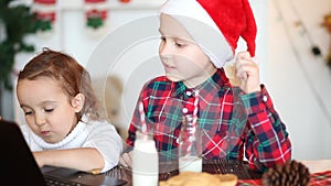 Funny kids girl and boy in Santa`s cap drinking Christmas milk, eating cookies and using a digital tablet laptop notebook. Online