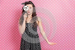 Funny kinky girl in headband and polkadot dress, hiding eye behind panda-like candy and making duck face.