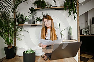 Funny lady with a laptop on the table and business papers in her hands, looking away with a pensive face on a white wall with