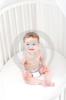 Funny laughing baby wearing a diaper sitting in a white round crib