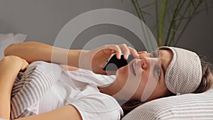 Funny laughing woman wearing blindfold lies in bed in fresh linen talking on mobile phone with her friend gossiping smiling and