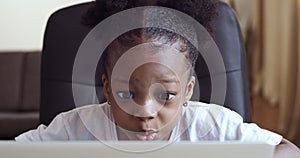 Funny little african girl looking at web camera or screen of laptop sitting at home, cute kid talking making online