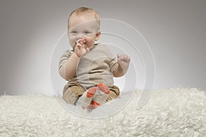 Funny little baby boy with fingers in the mouth, sitting on the white blanket, studio shot, isolated on grey background