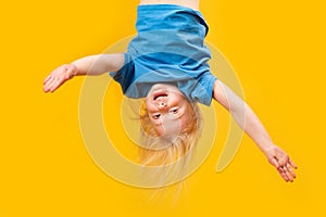 Funny little blonde girl upside-down on bright yellow background. Child topsy-turvy. Copy space