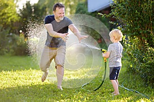 Funny little boy with his father playing with garden hose in sunny backyard. Preschooler child having fun with spray of water