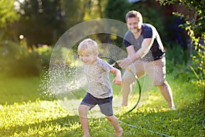 Funny little boy with his father playing with garden hose in sunny backyard. Preschooler child having fun with spray of water