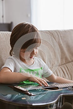 Funny little boy is learning to play the acoustic guitar at home, sitting on the couch and laughing.