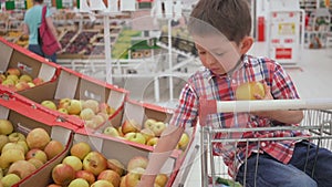 Funny little boy picking apple from the box sitting in the trolley, during family shopping in hypermarket