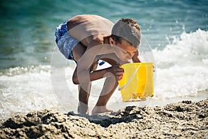 Funny little boy playing on beach with toy bucket.