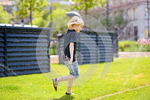 Funny little boy playing with lawn sprinkler in sunny city park. Elementary school child laughing, jumping and having fun with