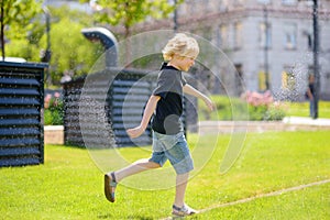 Funny little boy playing with lawn sprinkler in sunny city park. Elementary school child laughing, jumping and having fun with