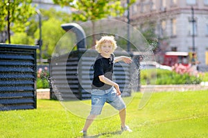 Funny little boy playing with lawn sprinkler in sunny city park. Elementary school child laughing, jumping and having fun with