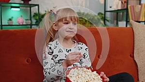 Funny little child girl watching comedy video film on tv, eating popcorn on comfortable sofa at home