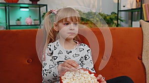 Funny little child girl watching comedy video film on tv, eating popcorn on comfortable sofa at home