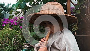 Funny little child in straw hat trying to play on flute with green garden on background. Happy boy playing on music instrument