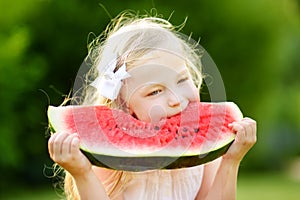 Funny little girl biting a slice of watermelon outdoors on warm and sunny summer day