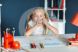 Funny little girl with blond hair sitting at white table and holding purple pencil