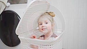 funny little girl blonde playing hide and seek in bathroom in laundry basket.