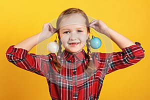 Funny little girl with christmas ball toys near the ears. Close-up of a child in a red dress on a yellow background
