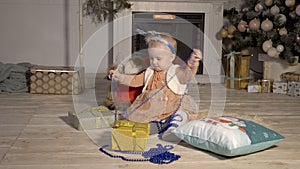 Funny little girl with Christmas decorations on her head playing with gifts, sitting on the floor near the fireplace and