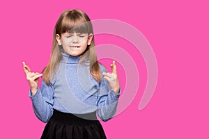 Funny little girl crossing fingers, hoping her wish come true, isolated on pink. Portrait of small girl praying, wishing good luck