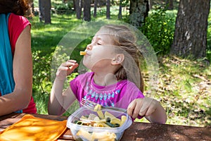 Funny little girl eating pasta from a plastic box next to her mother in a table picnic in countryside