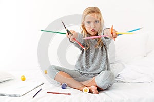 Funny little girl in gray pajamas playing with colorful pencils, looking at camera, sitting on bed