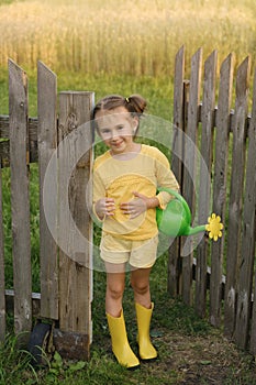 A funny little girl with a plastic watering can stands at the gate of an old wooden fence leading to the garden