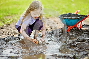 Funny little girl playing in a large wet mud puddle on sunny summer day. Child getting dirty while digging in muddy soil.