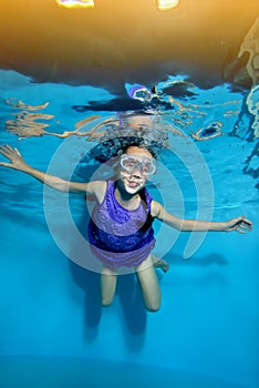 Funny little girl is swimming underwater in the pool on yellow light background in swimming glasses and purple dress.